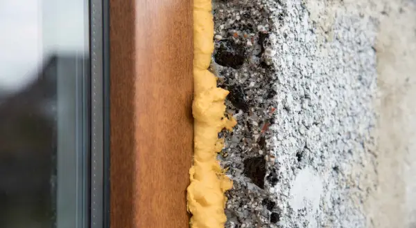 Dettaglio di un telaio di finestra in legno con schiuma poliuretanica espansa che sigilla lo spazio tra il telaio e il muro grezzo. Questo mostra come modellare la schiuma poliuretanica per un isolamento efficace.