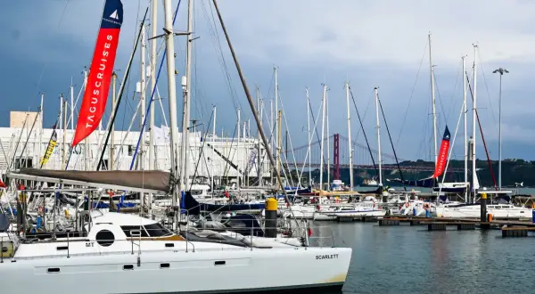 Una **barca lunga e sottile**, la Scarlett, è ormeggiata in un porto pieno di altre barche a vela. Sullo sfondo, un ponte rosso attraversa il fiume.