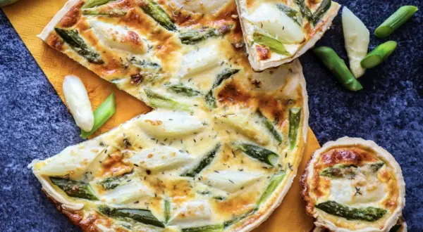 Herzhafte Quiche mit Spargel, goldbraun gebacken, auf einem orangen Tuch. Ein Stück ist angeschnitten, daneben liegen frische Spargelstücke.