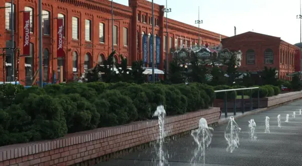 Piotrkowska Łódź: ceglane budynki Manufaktury, karuzela, fontanny i zielone krzewy tworzą letni klimat.
