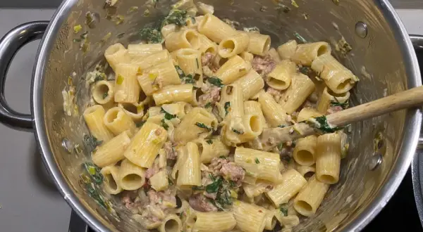Rigatoni mit cremiger Sauce, Fenchel und Salsiccia. Ein Holzlöffel rührt die köstliche Pasta in einem Topf.