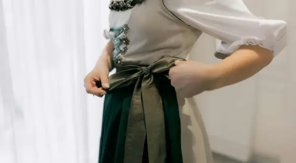 Eine Frau bindet die Schleife ihres Dirndls. Das Dirndl hat eine helle Weste mit Schnürung und eine dunkelgrüne Schürze.