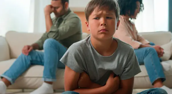 Bambino triste in primo piano, con i genitori separati seduti alle sue spalle, voltati l'uno dall'altra.