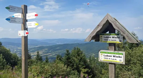 Znaki szlaku turystycznego na Luboń Wielki. W tle malowniczy krajobraz Beskidów i paralotniarz.