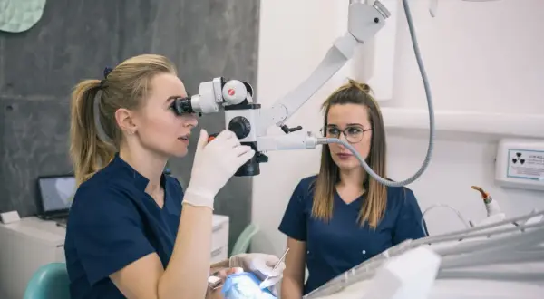 Dentystka z mikroskopem bada ząb pacjenta, by uniknąć chodzenia bez plomby. Pomaga jej asystentka.