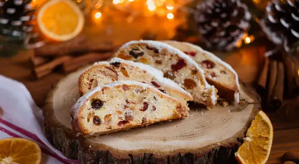 Tranches de stollen cake, un délice de Noël aux fruits secs et noix, saupoudré de sucre glace, sur une planche de bois, entouré de décorations festives.