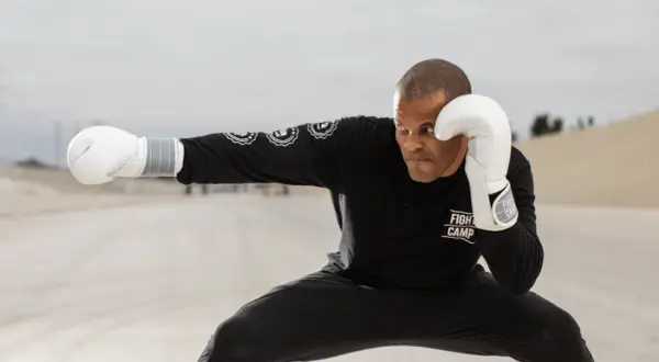 A boxer in a low crouching stance, wearing white gloves and a black "Fight Camp" shirt, throws a punch in an open, dusty landscape.