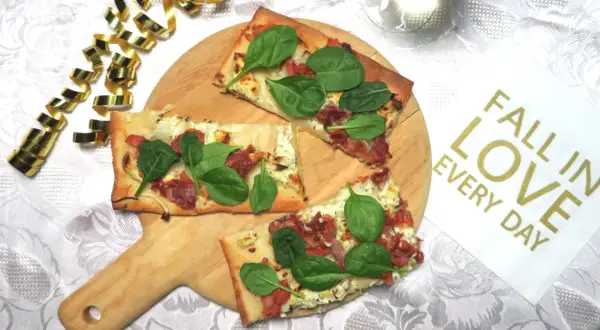 Prosciutto pizza med färsk spenat på en träskärbräda, omgiven av guldfärgad serpentin. En perfekt bit att "fall in love" med.