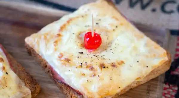Ein klassisches Toast Hawaii mit geschmolzenem Käse, Schinken und einer Kirsche, serviert auf einem Holzbrett.