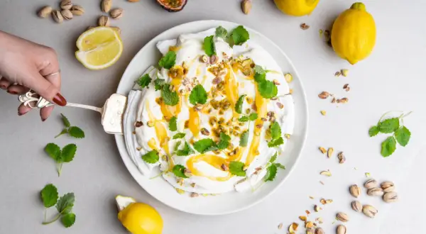 En hand skär en pavlova med lemon curd, toppad med passionsfrukt, pistagenötter och mynta.