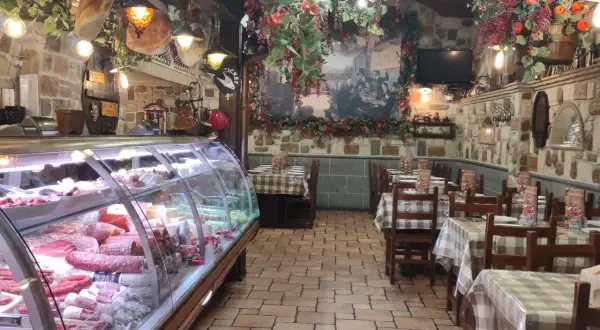 Wnętrze restauracji w stylu rustykalnym, z wiszącymi wędlinami i dekoracjami. W chłodni mnóstwo wyrobów, jak w najlepszym sklepie w Frascati.