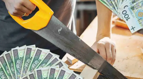 Cieśla tnie drewno piłą, a wokół leżą banknoty 100 zł. Zarobek rzemieślnika.
