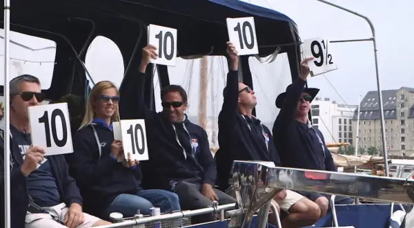 A group of diving judges hold up scorecards. Most show a perfect 10, but one judge holds up a 9.5.