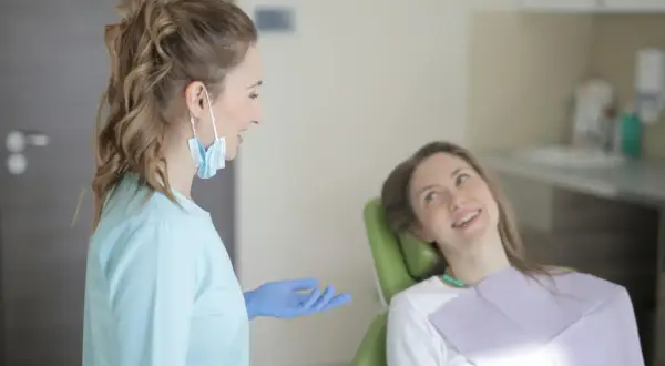 Dentystka w niebieskiej bluzie i rękawiczkach rozmawia z pacjentką. To obrazek pokazujący, jakie są studia stomatologiczne wymagania.
