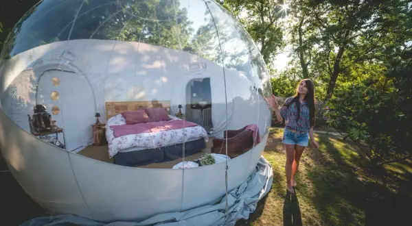 Une jeune femme admire sa **nuit bulle** confortable, un lit douillet dans une sphère transparente au milieu de la nature.