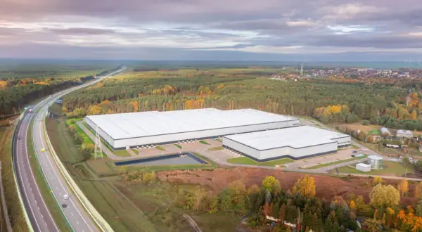 Nowoczesne, rozległe budynki firm magazynowych otoczone jesiennym lasem, w pobliżu autostrady.