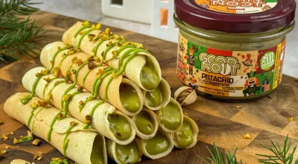 Stosik chrupiących rurek z kremem pistacjowym, polanych zielonym lukrem i posypanych orzeszkami. Obok słoik kremu i łuskane pistacje.
