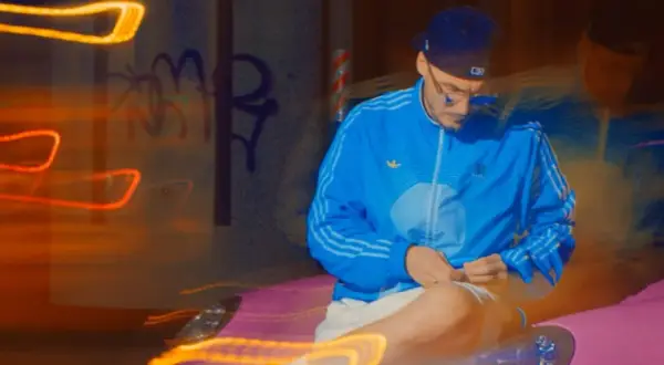 Mężczyzna w niebieskiej kurtce i czapce siedzi na tle nocnego miasta z neonowymi światłami.