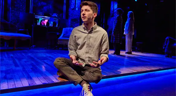 Andrew Barth Feldman sitzt im Schneidersitz auf einer Bühne, die in blaues Licht getaucht ist. Zwei Frauen stehen im Hintergrund.