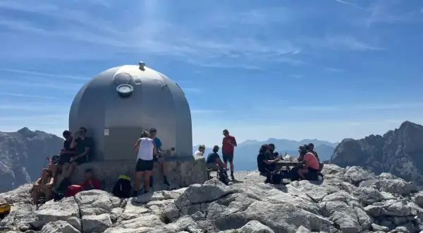 Gente descansa junto a un refugio en un viaje a Picos de Europa. El sol brilla en un cielo azul con nubes.