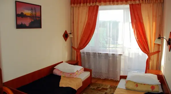 Pokój w sanatorium Plon Polańczyk. Dwa łóżka, okno z pomarańczowymi zasłonami, obraz na ścianie.