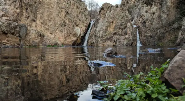 Cascadas gemelas caen entre rocas escarpadas en el embalse de Pedrezuela, reflejándose en el agua tranquila.