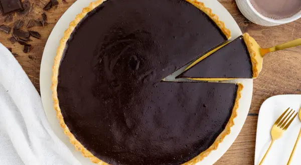 Ein Stück vom originalen Tarte au Chocolat Rezept wird mit einem goldenen Tortenheber herausgehoben. Daneben liegen Schokoladenstücke und eine Tasse Kakao.