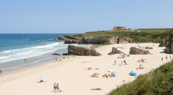 Gente disfrutando del sol y el mar en la playa Longara Barreiros, con formaciones rocosas y una casa amarilla al fondo.