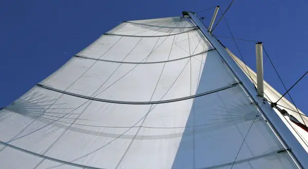 Vela bianca randa full batten spiegata al vento, con stecche orizzontali e cavi che salgono verso il cielo azzurro.