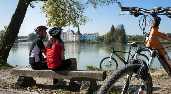 Para rowerzystów odpoczywa nad rzeką, podziwiając malownicze **trasy rowerowe Austrii** i pałac w oddali.