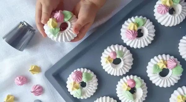 Dziecko ozdabia okrągłe mini bezy kolorowymi kleksami z kremu. Obok leży końcówka do szprycowania.