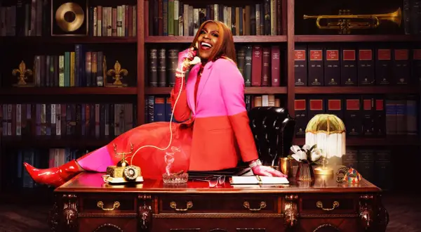 Big Freedia, in a vibrant pink and orange suit, sits at a desk, talking on a vintage phone. Her success is evident in the opulent office, hinting at her impressive big Freedia net worth.