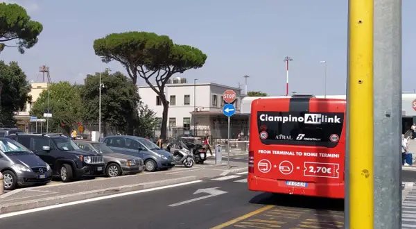 Czerwony autobus Ciampino Airlink czeka na pasażerów. Z tyłu widnieje napis "FROM TERMINAL TO ROME TERMINI" i cena 2,70€. W tle parking z samochodami i budynek lotniska.
