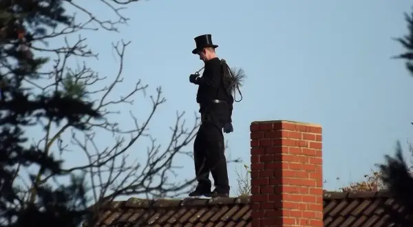Ein Schornsteinfeger in Zylinder und schwarzer Kleidung steht auf einem Dach. Er trägt seine Bürste und Kette bei sich. Man fragt sich, wie oft kommt der Schornsteinfeger wohl?