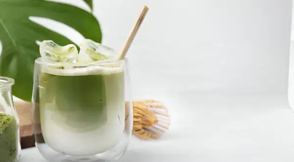Un verre de thé matcha glacé, parfait pour savoir si on peut boire le thé matcha froid. Une feuille de monstera et un fouet à matcha complètent la scène.