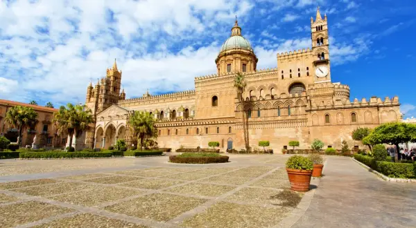 La maestosa cattedrale di Palermo si erge sotto un cielo azzurro con nuvole bianche, con la sua architettura imponente e il suo cortile lastricato.