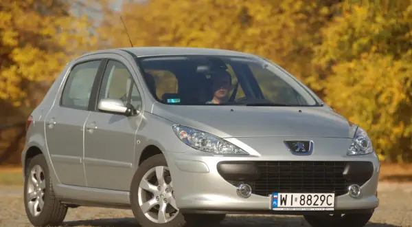 Srebrny Peugeot 307 z widocznymi żarówkami reflektorów, zaparkowany na tle jesiennych drzew.
