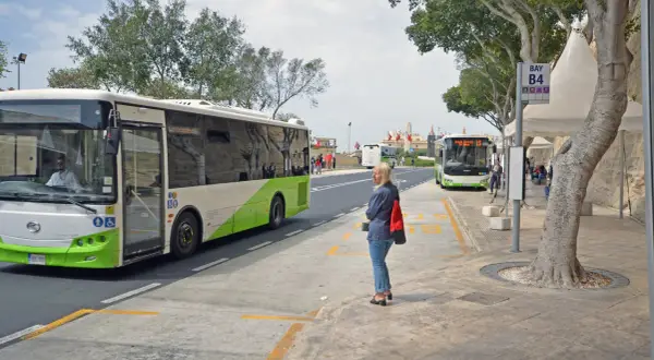 Kobieta czeka na autobus na przystanku przy lotnisku na Malcie. W tle widać inne autobusy i budynki.