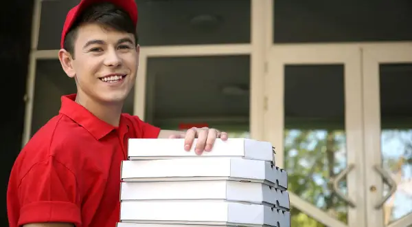 Uśmiechnięty pracownik młodociany w czerwonej koszulce i czapce niesie stos pudełek z pizzą.
