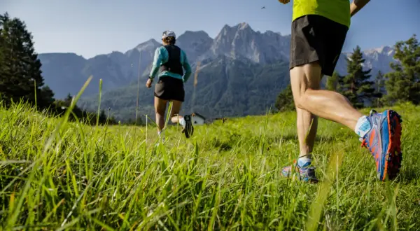 Zwei Läufer genießen das Zugspitze Laufen auf einer grünen Wiese vor majestätischen Bergen.
