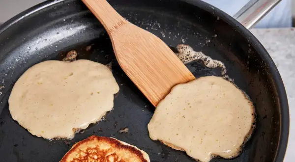 Buttermilch pancakes braten in der Pfanne. Ein Holzspatel wendet einen goldbraunen Pancake.