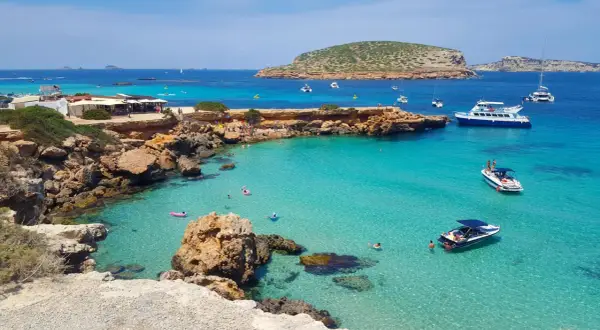 Plage idyllique à Ibiza avec eaux turquoise, rochers, bateaux et baigneurs. Parfait pour savoir où aller à Ibiza.