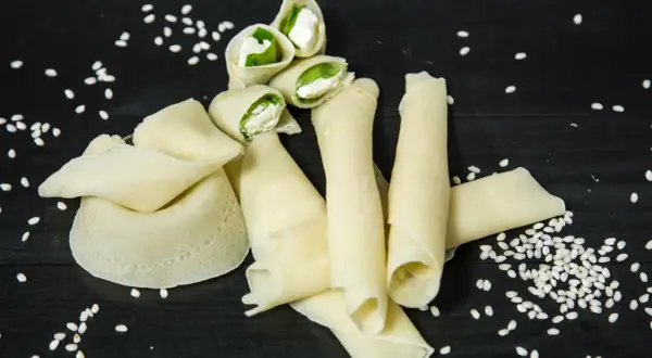 Rotolini di crepes con farina di riso, ripieni di formaggio fresco e basilico, sparsi su un tavolo scuro con semi di sesamo.