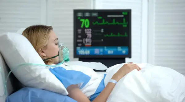 Kobieta w masce tlenowej leży w szpitalnym łóżku, monitor pokazuje parametry życiowe. Jej stan budzi niepokój, być może to rzucawka.