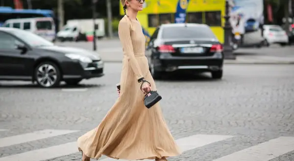 Elegancka, beżowa sukienka maxi, idealna na miejskie stylizacje. Kobieta w okularach przeciwsłonecznych i małej czarnej torebce przechodzi przez pasy.