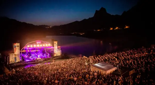 Gran concierto nocturno en el Pirineos Sur. Un mar de gente disfruta de la música bajo un cielo estrellado y montañas.