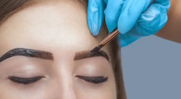 Stylist w niebieskiej rękawiczce precyzyjnie nakłada hennę na brwi, tworząc efekt brwi pudrowych.
