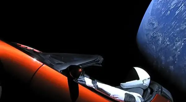 Pomarańczowy Tesla Roadster z manekinem w skafandrze kosmicznym dryfuje w kosmosie obok Ziemi.