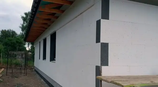 Dom w trakcie budowy, widoczna biała izolacja ścian zewnętrznych z szarymi narożnikami. Dach z drewnianymi krokwiami.