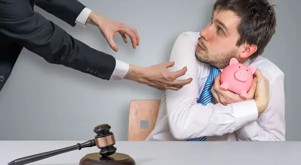 Młody mężczyzna kurczowo trzyma skarbonkę, gdy ręce w garniturze wyciągają się ku niemu. Młotek sędziowski leży na stole, symbolizując upadłość konsumencką.